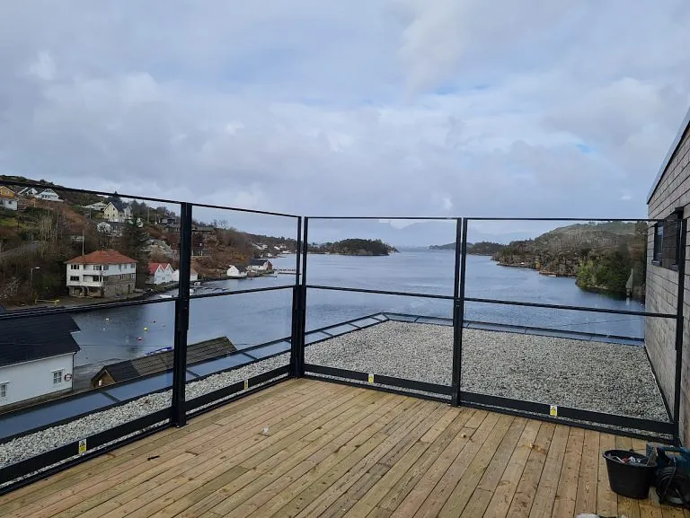 Terrasse med høyt svart glassrekkverk og utsikt over fjord og hus.
